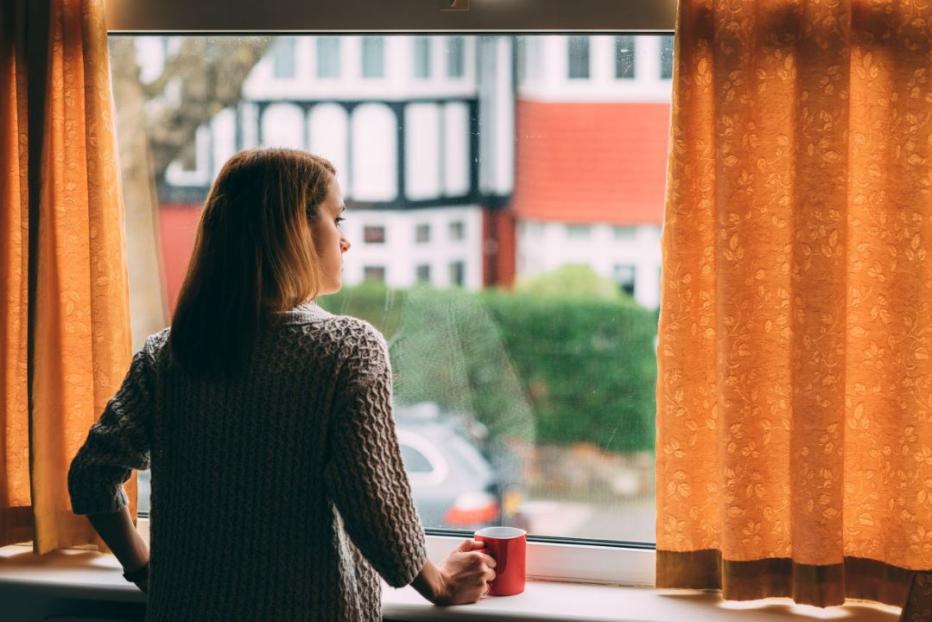 Stock image of woman in a beige sweater looking out a window draped with gold curtains